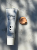 A tube of Verso Skincare Daily Glow cream with retinol and vitamin C, placed on a light marble surface with natural leaf shadows, next to a dollop of the beige-colored cream.